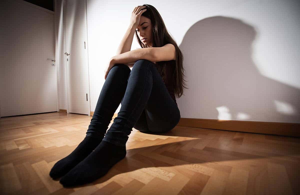 an anxious female teen sits on the floor against the white wall of her room