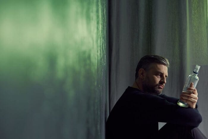 a bearded man sits on the floor looking at the empty glass of alcohol bottle on his hand