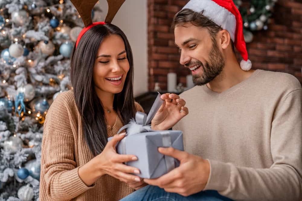 A couple exchanging a blue gift box in front of a Christmas tree