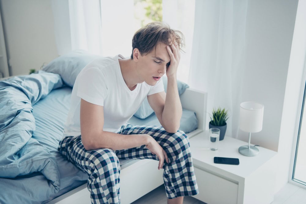 A man sitting on the bed wearing white shirt and blue pajamas, holding his forehead due to a hangover