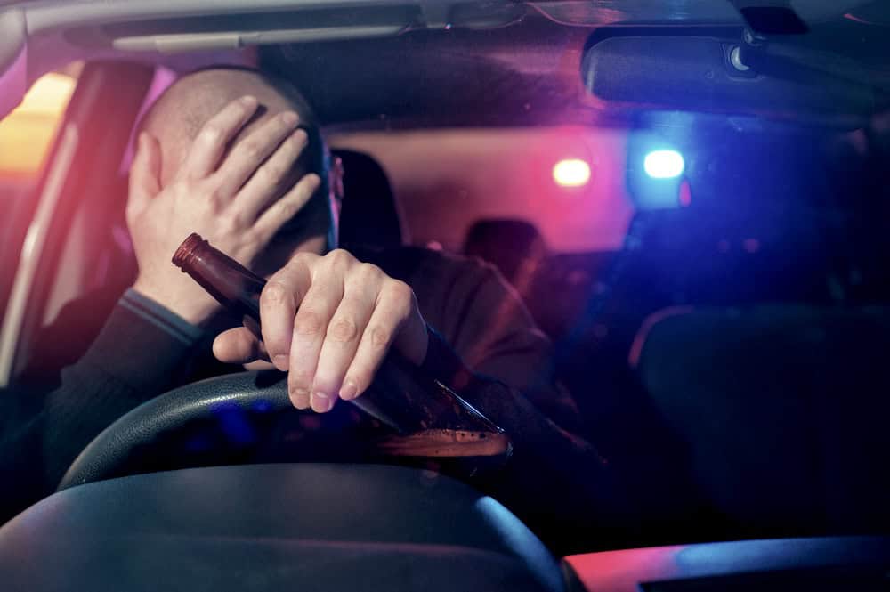 Man behind the wheel of a car holding a beer bottle while police car lights are seen in the background