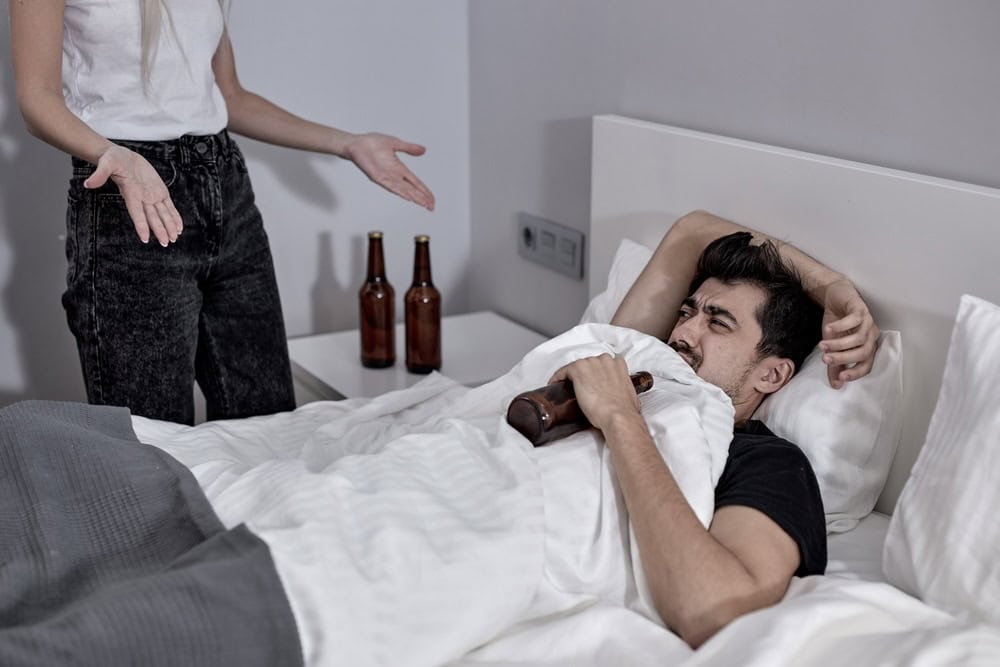 Husband denying his addiction lying on a bed with alcohol bottle 